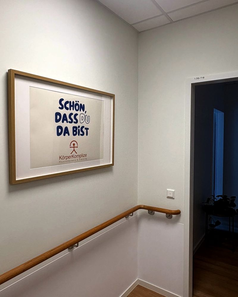 Treppenhaus der Praxis: ein gerahmtes Plakat mit dem Schriftzug „Schön, dass du da bist“ und dem Logo KörperKomplize, daneben ein Handlauf und die offene Tür zur ersten Etage.
