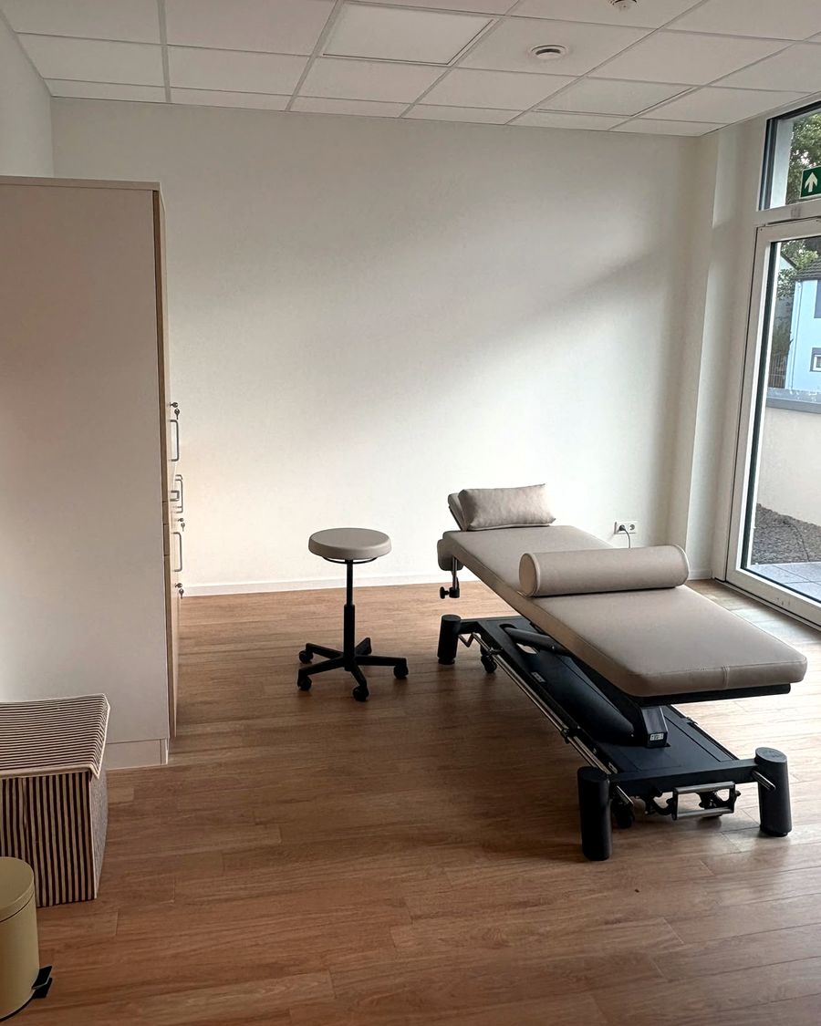 Heller Behandlungsraum mit Therapieliege, Nackenrolle und Kissen vor einer weißen Wand, daneben ein Rollhocker, rechts ein bodentiefes Fenster.