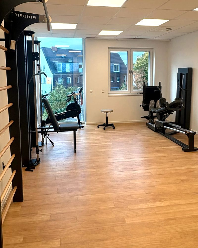 Trainingsfläche mit Sprossenwand, Seilzug, Trainingsbank, Ergometer am bodentiefen Fenster und einer Beinpresse an der rechten Wand.
